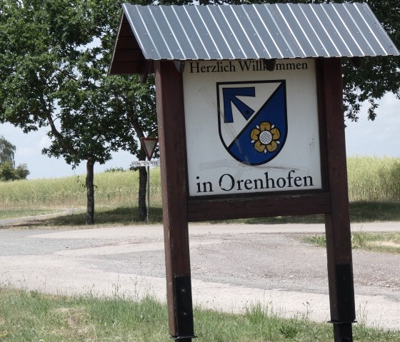 Panneau de la localité d'Orenhofen avec armoiries, entouré d'arbres et de champs. Un chemin traverse le paysage rural., © TI Bitburger Land