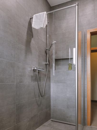 Eine moderne Duschkabine aus Glas mit grauen Fliesen. Ein Handtuch hängt an der Wand und es gibt eine kleine Ablage für Pflegeprodukte.