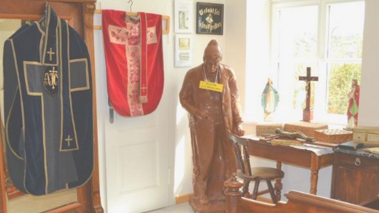 Ein traditionelles Zimmer mit religiösen Gegenständen. An der Wand hängen Gewänder und eine Statue steht an einem Tisch.