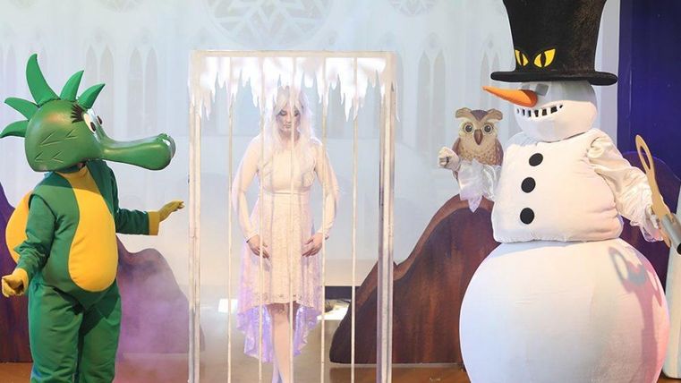 Eine bunte Theateraufführung mit einem Drachen, einem Schneemann und einer gefangenen Frau in einem eisigen Käfig. Die Charaktere tragen auffällige Kostüme und stehen auf einer festlichen Bühne.