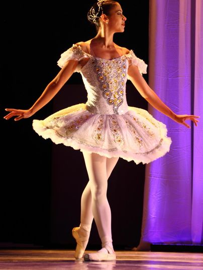 Eine Balletttänzerin in einem eleganten Kostüm steht auf der Bühne. Ihr Outfit ist weiß mit glitzernden Verzierungen und die Beleuchtung schafft eine festliche Atmosphäre.
