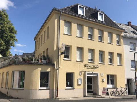A yellow building with three floors and shutters. The facade has plants on the balcony and bicycles standing in front.