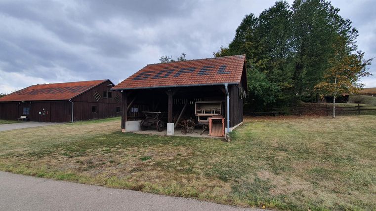 A rural building with a red roof stands next to a meadow. In the background, trees can be seen and the sky is cloudy.