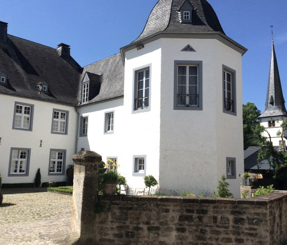 Wit kasteel met grijs dak en toren, omgeven door tuin en stenen muur, onder een blauwe hemel., © Fellmann / TI Bitburger Land