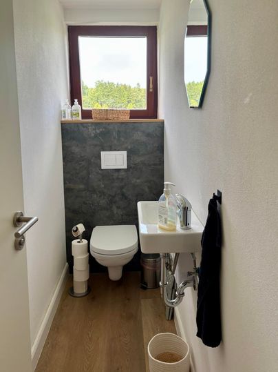 Une petite toilette moderne avec un lavabo blanc et une lunette de toilettes. La fenêtre laisse entrer beaucoup de lumière et montre un paysage verdoyant à l'extérieur.