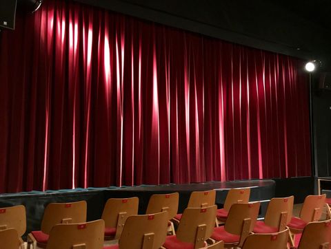 A red curtain on the stage of a theater. In front of the stage, there are empty wooden chairs.