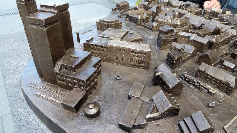 Eine detaillierte Bronze-Tafel zeigt ein Stadtmodell mit Gebäuden und Straßen. Im Hintergrund ist ein hoher Wolkenkratzer zu sehen.