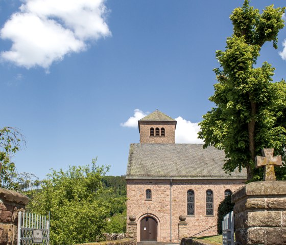 L'église St. Maximin à Kyllburg, entourée d'arbres, sous un ciel bleu clair. Au premier plan, on peut voir des croix en pierre., © TI Bitburger Land