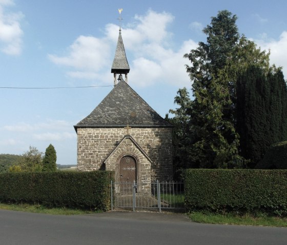 Chapelle en pierre avec toit pointu et girouette, entourée d'arbres et de haies, sur fond de ciel bleu., © Conny Meier