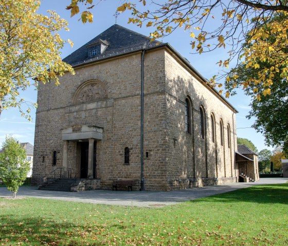 Die Kirche St. Hubertus in Wolsfeld, umgeben von herbstlichen Bäumen, steht in einem grünen Park. Das Gebäude ist aus Stein mit einem Satteldach., © TI Bitburger Land