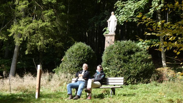 Zwei Personen sitzen auf einer Bank vor einer Mariensäule im Wald.
