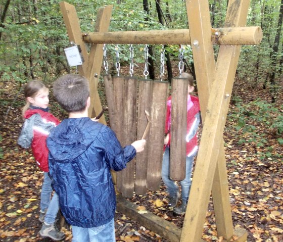 Children play at a wooden sound station in the forest. They are wearing rain jackets and standing on a leaf-covered floor., © Tourist-Information Bitburger Land