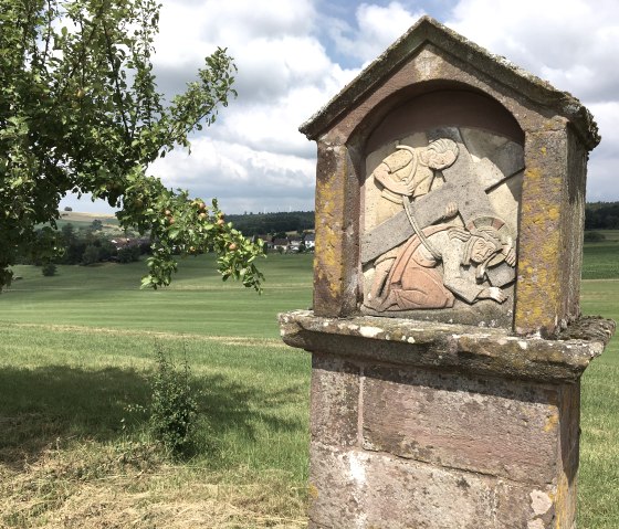 Steinrelief eines Kreuzwegs in ländlicher Landschaft. Ein Apfelbaum mit reifen Früchten steht im Vordergrund, grüne Felder und ein Dorf im Hintergrund., © TI Bitburger Land