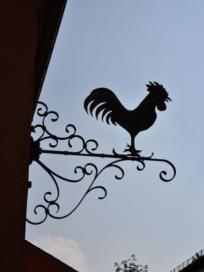 Scherenschnitt eines Hahns auf einem kunstvollen Metallschild vor blauem Himmel.