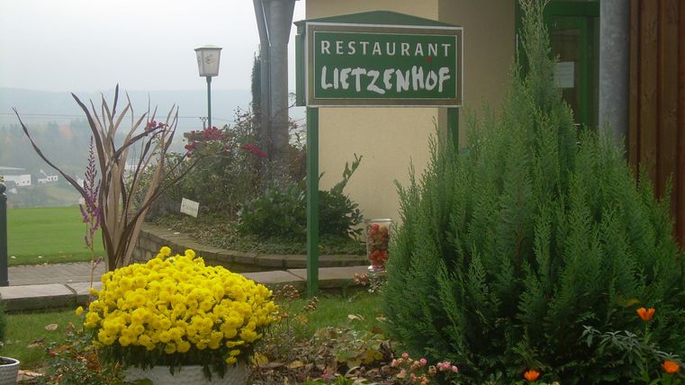 Ein Restaurant mit dem Namen "Lietzenhof" ist sichtbar, umgeben von einem blühenden Garten. Bunte Blumen und Pflanzen schmücken den Eingangsbereich.