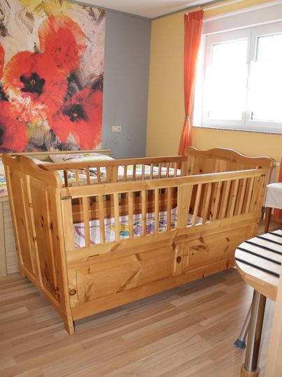 Ein gemütliches Kinderbett aus Holz steht in einem hellen Raum. An der Wand hängen lebhafte Bilder und die Fenster lassen viel Tageslicht herein.