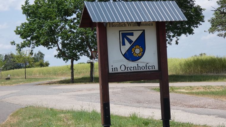 Ortsschild von Orenhofen mit Wappen, umgeben von Bäumen und Feldern.