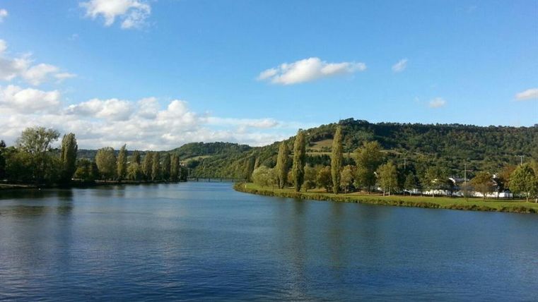 Une rivière tranquille avec des vagues douces et des rives verdoyantes. En arrière-plan, des collines et un ciel bleu avec quelques nuages sont visibles.