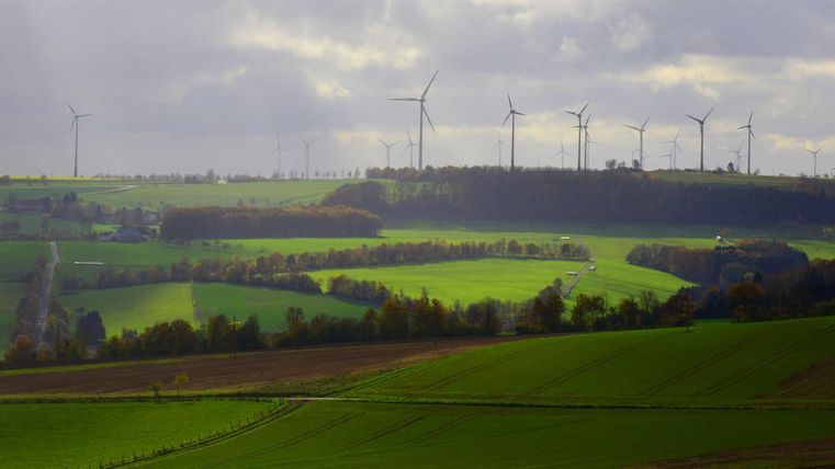 Eine grüne Landschaft mit sanften Hügeln und Windkraftanlagen im Hintergrund. Der Himmel ist bewölkt und es gibt sanfte Lichtstrahlen.