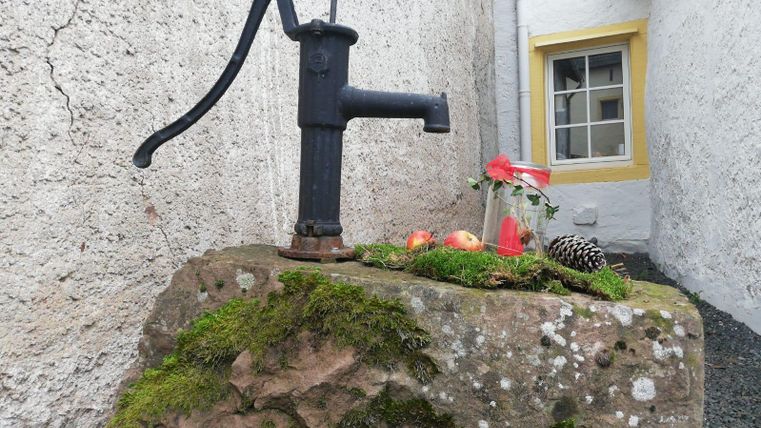 An old-fashioned water pump stands on a moss-covered stone. Next to it lie some apples and a pine cone.
