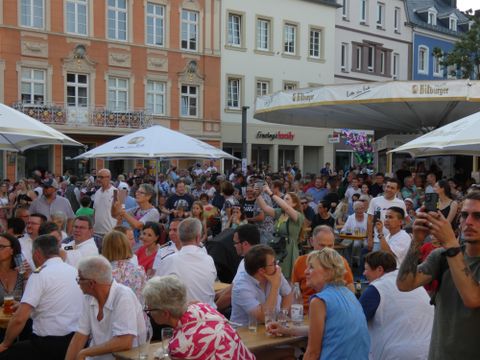 A lively square situation with many people sitting at tables and celebrating with each other. Some guests are holding drinks in their hands, and the atmosphere feels cheerful and sociable.