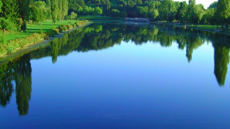 Ein ruhiger See, umgeben von üppigem Grün und Bäumen. Die spiegelnde Wasseroberfläche reflektiert die umliegende Landschaft.