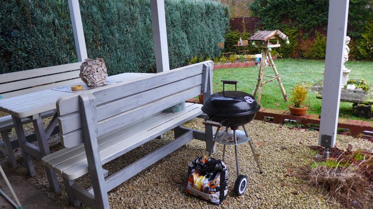 A cozy terrace with a wooden table and benches. There is a grill on it, and the garden is well-kept.