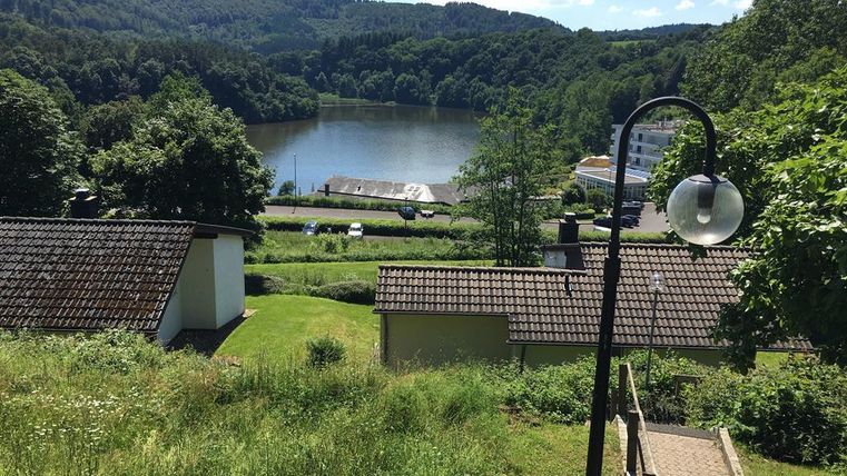 Eine malerische Landschaft mit einem ruhigen See und üppigem Grün. Im Hintergrund sind Hügel und einige Gebäude zu sehen.