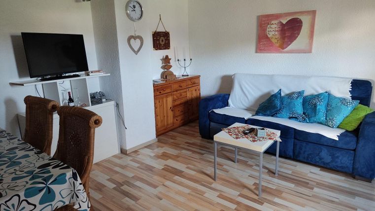 Ein gemütliches Wohnzimmer mit einem blauen Sofa, bunten Kissen und einem Tisch. In der Ecke steht ein Fernseher und es gibt einen kleinen Essbereich mit einer blumigen Tischdecke.