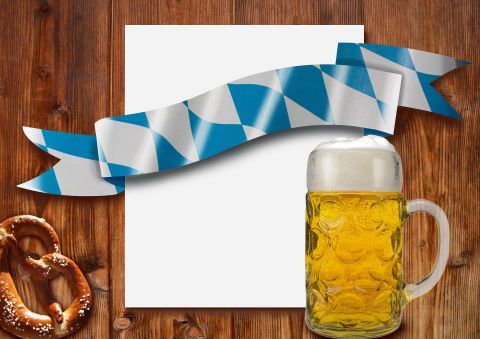 Een bierpul met gouden bier staat naast een pretzel op een houten tafel. Op de achtergrond ligt een leeg vel en een decoratieve blauwe en witte strik.