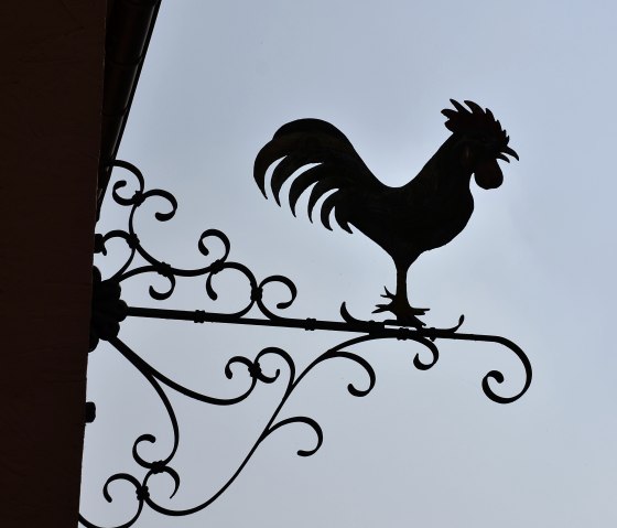 Scherenschnitt eines Hahns auf einem kunstvoll verzierten Metallschild vor klarem Himmel., © TI Bitburger Land