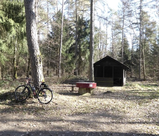 Un vélo est adossé à un arbre près d'un banc dans la forêt. En arrière-plan se trouve une petite cabane en bois., © Eifelverein Ortsgruppe Speicher