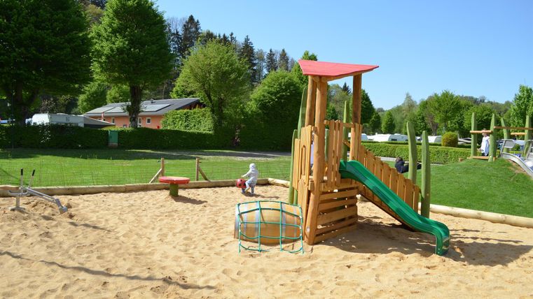 Ein schöner Spielplatz mit einer Rutsche und Sandkasten. Umgeben von grünen Bäumen und Wiesen.