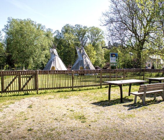 Een speeltuin met twee tipi's achter een houten hek, omringd door bomen. Op de voorgrond staan tafels en banken op een grindvloer., © Tourist-Information Bitburger Land_M. Mayer