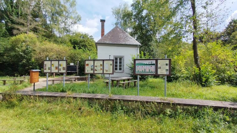 Ein kleines weißes Gebäude mit Schautafeln und Bänken davor, umgeben von Bäumen und Gras.