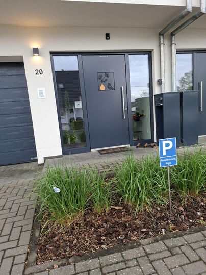 Une maison moderne avec une porte d'entrée grise et un panneau de stationnement devant. Au premier plan, des plantes vertes poussent le long du chemin.