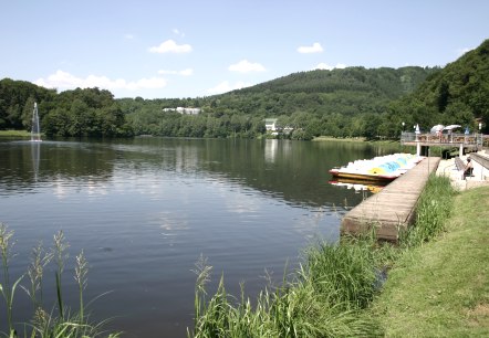Le lac de barrage de Bitburg aux eaux calmes, entouré de collines verdoyantes. Des pédalos sont amarrés au ponton, des gens se détendent sur la rive. Une belle journée d'été., © TI Bitburger Land