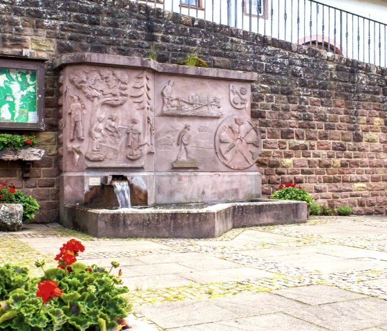 Ein steinerner Dorfbrunnen mit Reliefs und fließendem Wasser, umgeben von roten Blumen und einer Infotafel an einer Steinmauer., © TI Bitburger Land - M. Mayer