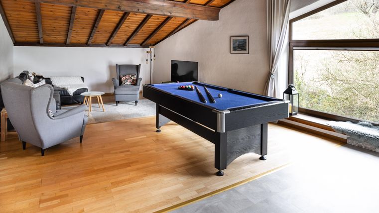 Ein stilvolles Wohnzimmer mit einem Billardtisch und einer gemütlichen Sitzecke. Große Fenster sorgen für viel natürliches Licht und einen Blick nach draußen.
