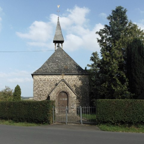 Stone chapel with pointed roof and weathercock, surrounded by trees and hedges, against a blue sky., © Conny Meier