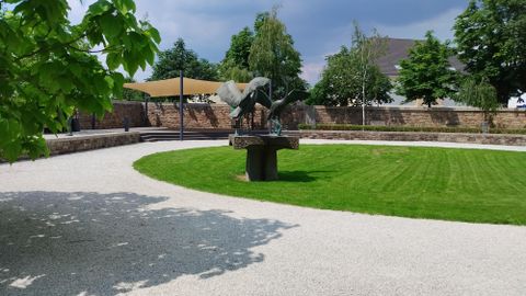 Un jardin soigné avec de l'herbe et un chemin de gravier. Au milieu se trouve une sculpture entourée d'arbres et de buissons.
