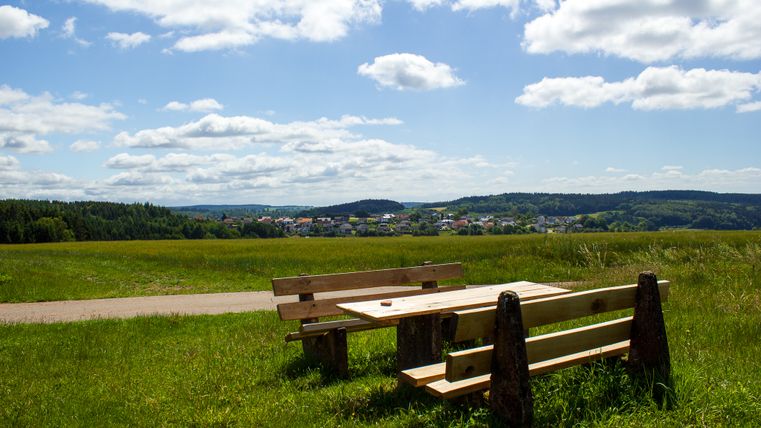 Holztisch und Bänke auf einer Wiese mit Blick auf Neidenbach und bewaldete Hügel im Hintergrund.