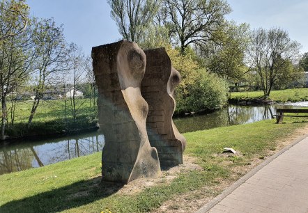 Stenen sculptuur aan de oever van de rivier, omgeven door groen landschap en bomen. Een heldere hemel en een pad zijn ook zichtbaar., &copy; TI Bitburger Land, Steffi Wagner