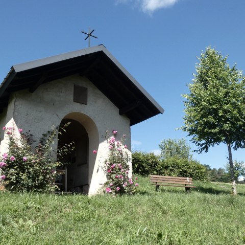 Kleine Kapelle im Grünen, umgeben von blühenden Rosen. Eine Bank steht daneben, ein Baum spendet Schatten. Blauer Himmel im Hintergrund., © Tourist-Information Bitburger Land, Melanie Salzburger