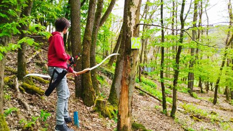 Ein Junge steht im Wald und zielt mit einem Bogen auf einen Baum. Um ihn herum sind grüne Bäume und frisches Laub.