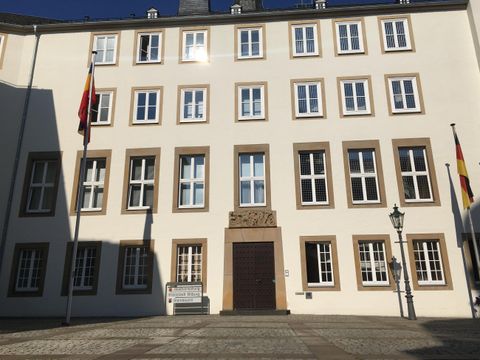 Ein historisches Gebäude mit mehreren Fenstern und zwei Fahnen. Die Fassade ist hell und klar, umgeben von einem Platz.