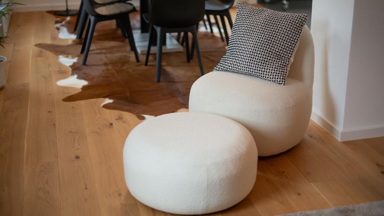 Een moderne ruimte met een comfortabele stoel en een ronde pouf in heldere kleuren. De vloer is van hout en er staat een eettafel op de achtergrond.