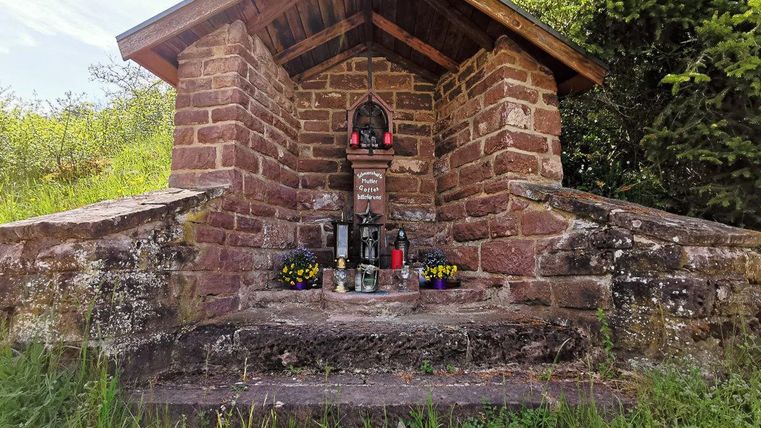 Ein steinernes Wegekreuz mit Dach, umgeben von Pflanzen, mit Blumen und Kerzen dekoriert.