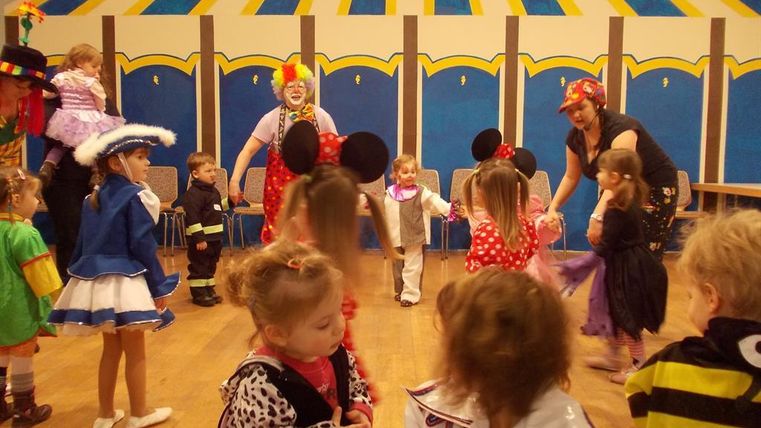 A cheerful children's masquerade with many dressed-up kids in colorful costumes. In the background, clowns and adults join in while the children dance and laugh.