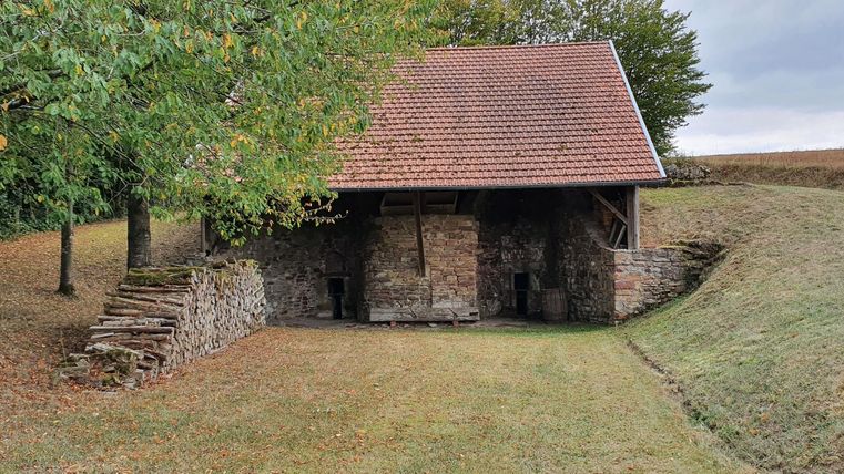 A small, traditional stone house with a red roof stands in a green landscape. Trees surround the building, and the sky is overcast.